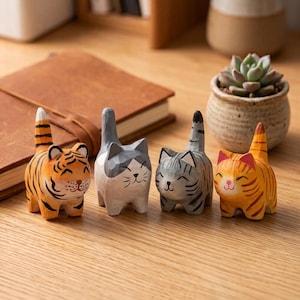 Puede incluir: Cuatro pequeñas figuras de animales de madera talladas a mano dispuestas en fila. El conjunto incluye un tigre con rayas naranjas y negras, un gato gris y blanco, un gato atigrado gris y un gato atigrado naranja. Cada figura sonríe.