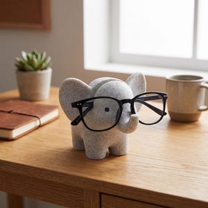 May include: A light grey elephant-shaped eyeglass holder with a speckled texture. The elephant has large ears and a trunk that supports a pair of black-rimmed eyeglasses. The object is made of a smooth material and features a small dark eye.
