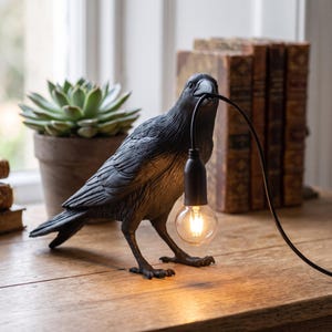 May include: A matte black raven-shaped table lamp featuring a textured feather design. The bird holds a black electrical cord in its beak, which suspends a single clear glass light bulb with a visible filament, casting a warm glow downward.