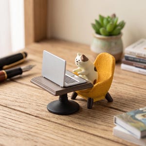 May include: A small figurine of a cream and brown cat sitting in a textured yellow armchair at a brown table with a silver laptop. The cat has simple painted features including black eyes, a small nose, and whiskers, facing the open laptop screen.