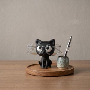 May include: A matte black cat-shaped figurine with large white eyes wearing clear-framed glasses, positioned on a round wooden tray next to a small speckled ceramic cup holding a silver fountain pen with a clear barrel and black ink.
