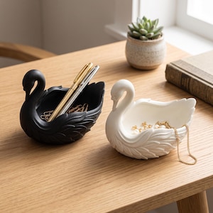 May include: Two swan-shaped bowls, one matte black and one matte white, featuring sculpted feather details. The black bowl holds two metallic pens and several paper clips, while the white bowl contains a pair of gold earrings and a gold chain necklace.