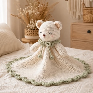 Peut inclure: Une couverture de sécurité en crochet couleur crème avec une tête d'ours aux yeux brodés, un nez marron et des joues roses. La couverture présente une bordure festonnée vert sauge et un lien assorti au cou avec deux petits pompons.