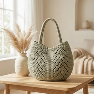 Puede incluir: Un bolso de mano de ganchillo en color verde salvia con una silueta estructurada y redondeada, un diseño de punto en espiga y dos asas integradas. El bolso está hecho de hilo grueso, mostrando una textura de punto calado.