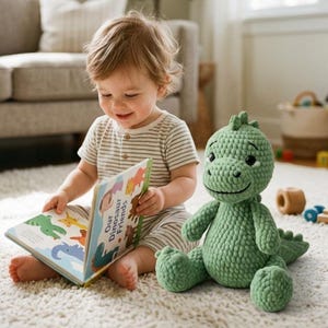 Può includere: Un peluche a forma di dinosauro lavorato all'uncinetto in verde chiaro con una finitura in filato morbido e testurizzato, dotato di testa arrotondata, braccia piccole e coda, seduto accanto a un libro per bambini intitolato Our Dinosaur Friends.