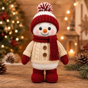May include: A crocheted snowman doll wearing a white and red pom-pom hat, a red scarf, a beige cable-knit sweater with two brown buttons, red mittens, and red boots. The snowman has a small orange carrot nose, black eyes, and pink cheeks.