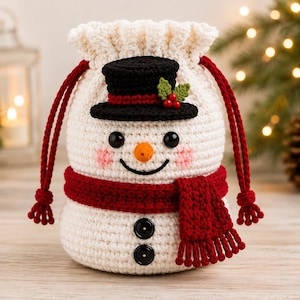 May include: A cream-coloured crochet drawstring pouch shaped like a snowman. It features a black top hat with red trim and holly, a bright orange nose, black button eyes, pink cheeks, a red scarf, and two black buttons on the lower body.