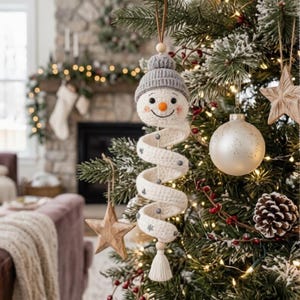 May include: A crocheted snowman ornament featuring a grey ribbed beanie, a white spiral body with small grey accents, and a bottom tassel. The snowman has a round face with a small orange nose, black eyes, and a stitched smile, hanging by a cord.