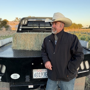 May include: A light-colored cowboy hat with a woven pattern on the crown, paired with a black zip-up jacket and blue denim jeans. The jacket features a small white logo on the chest. The attire is worn by a person with a beard and mustache.