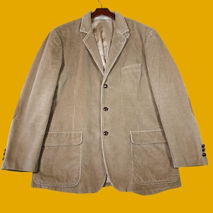 May include: A tan corduroy blazer featuring a notched lapel, three dark brown buttons down the front, and two large patch pockets at the waist. The sleeves have three matching buttons at the cuffs, and the interior is lined with a smooth fabric.