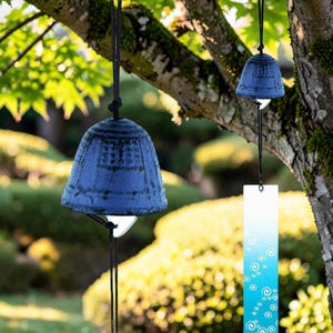 May include: Two dark blue cast iron wind chimes hanging from a tree branch. Each bell features a textured surface and a rectangular wind catcher. The wind catchers are white and blue with a repeating spiral pattern printed on the surface.