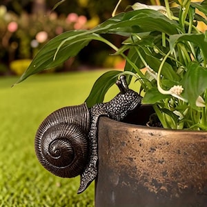 May include: A dark metallic snail figurine with a textured, spiralled shell and two antennae, designed to hook onto the rim of a cylindrical bronze-toned planter containing a green leafy plant with white flowers.