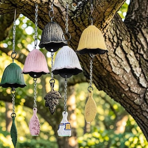 May include: A collection of six hanging metal wind chimes in green, pink, black, white, and yellow. Each bell features a fluted design and hangs from a silver chain with a decorative metal leaf or paddle-shaped pendant attached at the bottom.