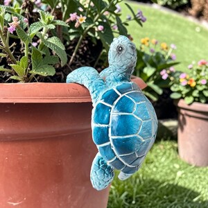 May include: A small, blue sea turtle figurine with a textured shell and detailed flippers, designed to hook onto the rim of a terracotta-coloured plant pot. The turtle has a glossy finish and is positioned as if climbing over the edge of the pot.
