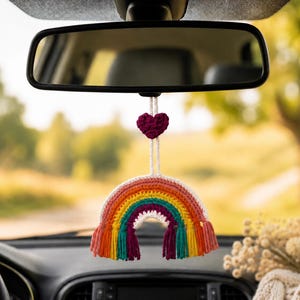 Peut inclure: Un accessoire de voiture arc-en-ciel en crochet suspendu à un rétroviseur. L'arc-en-ciel présente des arches concentriques en rose, orange, jaune, vert et violet, terminées par des pompons en fil assortis et un petit cœur en crochet violet.