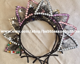 May include: Six black headband cat ears with jeweled embellishments. The ears are decorated with rhinestones, pearls, and lace. The headband is flat-backed and has a black elastic band.