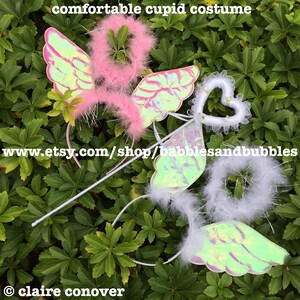 May include: Two sets of iridescent angel wings with white and pink feather halos, and a white heart-shaped wand with a white lace trim. The wings are attached to headbands and the wand has a silver stick.