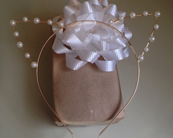 May include: A gold headband with pearl accents shaped into cat ears. The headband is on top of a brown gift box with a white satin bow.