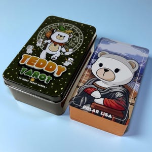 May include: A rectangular dark green metal tin labeled Teddy Tarot with 79 Teddy Tarot Cards text and a white bear illustration, alongside a deck of cards featuring a white bear character titled Polar Lisa with orange-colored card edges.