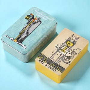 Op de afbeelding: Een lichtblauw metalen blik met de illustratie van de tarotkaart de Kluizenaar en de tekst Borderless-Waite Tarot Deck, naast een stapel kaarten met een beige achterkant met de illustratie van de Aas van Kelken en de tekst Ace of Cups.