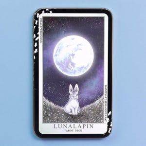 May include: A rectangular metal tin featuring the Lunalapin Tarot Deck. The lid displays a white rabbit sitting in a field under a large, glowing full moon against a dark purple starry sky. The text reads Lunalapin Tarot Deck and Mali Publishing.