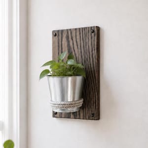 May include: A wall-mounted planter featuring a rectangular dark stained wood board with visible grain. A silver metal bucket is attached to the board, wrapped with a thick white rope detail. A green leafy plant grows inside the metal bucket.
