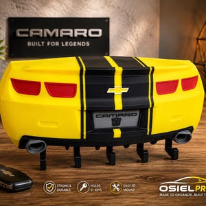 May include: A yellow and black wall-mounted key holder shaped like the rear of a Chevrolet Camaro. It features red taillights, a central Chevrolet emblem, a Camaro license plate with a Transformers logo, and five black hooks for hanging keys.