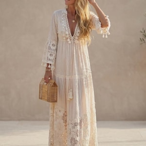 May include: A floor-length cream-colored maxi dress featuring a deep V-neckline, three-quarter length sleeves, and intricate lace paneling. The garment includes tassel accents along the neckline and hem, paired with a structured wooden handbag.