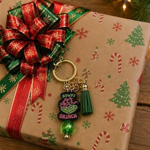 May include: A gold-toned metal keychain featuring a black rectangular charm with a green Grinch character wearing a pink cowgirl hat, the text HOWDY GRINCH, a green tassel, and a green bead, attached to a gift box with a red and green bow.