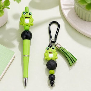 May include: A light green pen topped with a cartoon frog figure and a smooth black bead, alongside a keychain featuring a matching frog figure, black beads, a textured black bead, a green suede tassel, and a black metal carabiner clip.