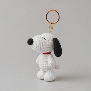 Puede incluir: Un llavero de perro tejido a ganchillo en color blanco con una nariz negra y una oreja negra. La figura lleva un fino collar rojo alrededor del cuello y está unida a una anilla de metal dorado mediante una cadena corta. Se aprecia la textura.