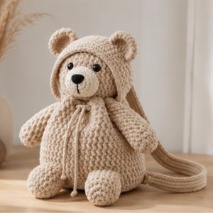 Könnte beinhalten: Ein beigefarbener Häkelrucksack in Form eines Teddybären mit Kapuze, runden Ohren, schwarzen Kunststoffaugen und einer schwarz gestickten Nase. Die Tasche verfügt über einen Kordelzugverschluss und zwei lange Träger aus dickem Garn.