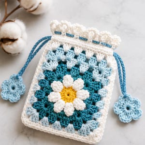 May include: A rectangular crochet drawstring pouch featuring a central white and yellow daisy motif surrounded by layers of teal and light blue yarn. The top edge has a white scalloped border with blue drawstrings ending in small blue flower charms.