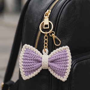 Può includere: Un portachiavi a forma di fiocco all'uncinetto con un centro color lavanda e un bordo color crema. Il fiocco è fissato a un anello in metallo dorato e a un moschettone, agganciato alla zip di uno zaino in pelle nera.