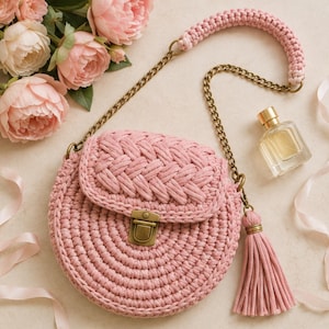 May include: A round, dusty pink crocheted handbag featuring a braided flap, a brass-toned metal push-lock closure, and a matching tassel. The bag includes a long chain strap with a thick, crocheted shoulder pad in the same pink cord material.