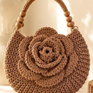 Könnte beinhalten: Eine runde, hellbraune Häkeltasche mit einem großen, dreidimensionalen rosenförmigen Design auf der Vorderseite. Die Tasche verfügt über einen einzelnen, dicken, geflochtenen Henkel, der an beiden Seiten mit drei glatten Holzperlen verziert ist.