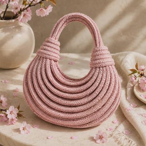 Può includere: Una borsa a mezzaluna realizzata in filato lavorato all'uncinetto di colore rosa chiaro. La borsa presenta un design strutturato e a coste con diversi strati concentrici che formano il corpo e un manico integrato annodato alle estremità.