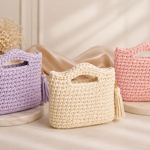 May include: Three small, rectangular crochet handbags with integrated handles. The bags are coloured lavender, cream, and light pink. Each bag features a matching decorative tassel attached to the side with a gold-toned metal clasp.