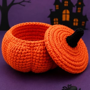 May include: A round, orange, crocheted pumpkin-shaped container with a removable lid. The lid features a small, black, crocheted stem at the center. The container has a textured, ribbed surface created by the crochet stitches, mimicking a pumpkin.