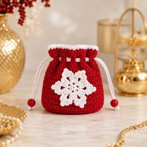 May include: A small, round, red crochet drawstring pouch featuring a white crocheted snowflake appliqué on the front. The pouch has a white drawstring cord with red beads at the ends and a white crocheted trim along the top opening edge.