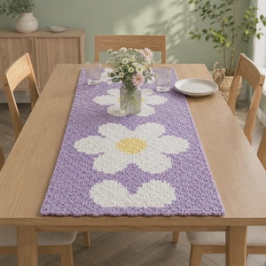 Könnte beinhalten: Ein rechteckiger Häkel-Tischläufer in hellem Violett mit einem sich wiederholenden Muster aus großen weißen Blumen mit gelben Mitten. Der Tischläufer liegt flach auf einem hölzernen Esstisch unter einer mit Blumen gefüllten Glasvase.