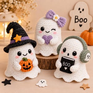 May include: Three white crocheted ghost figures. One wears a black witch hat with a yellow star and holds an orange pumpkin. Another has a purple bow with a white flower and holds a purple heart. The third wears green headphones and holds a black book labelled BOO.