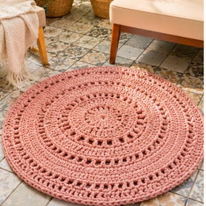 May include: A circular, dusty rose-colored rug featuring a complex, concentric crochet pattern with openwork stitches. The rug is crafted from thick, textured yarn, creating a repeating geometric design that radiates from the center outward.