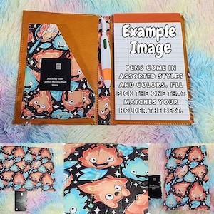 May include: A tan leather-effect notebook holder featuring a pocket with a black background and a colourful cartoon flame pattern. It includes a white pen, a lined notepad with the text Example Image, and a black card with white text.