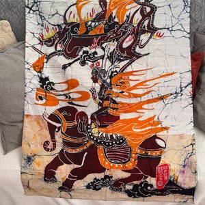 May include: A rectangular batik fabric wall hanging featuring a white background with a crackled texture. The design depicts a figure riding a dark brown elephant, surrounded by orange flames and a dark dragon-like creature with red accents.
