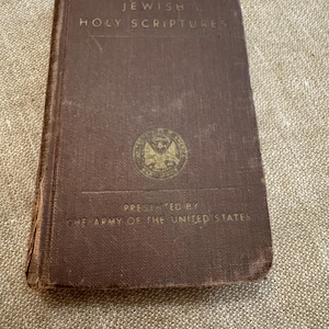 May include: A brown hardback book titled Jewish Holy Scriptures with a gold circular emblem in the centre. The cover features the text Presented by The Army of the United States printed in gold lettering below the emblem on the front cover.
