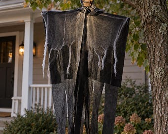 May include: A hanging Halloween decoration featuring a beige skull head with hollow eye sockets and a nose cavity, draped in layers of tattered, translucent black fabric that hangs down in long, uneven strips from a central hood structure.