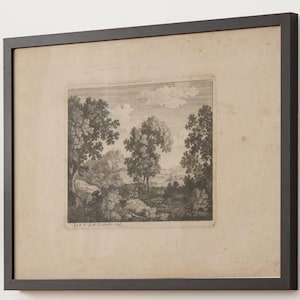 Puede incluir: Un grabado en blanco y negro enmarcado que representa un paisaje con árboles, rocas y un horizonte lejano bajo un cielo nublado. La obra está centrada sobre un paspartú beige claro en un marco negro fino. El texto dice Jan v. d. Velde fecit 1616.