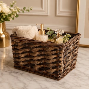 May include: A rectangular dark brown woven wicker basket containing a folded beige towel, a clear glass soap dispenser with a gold pump, a small glass perfume bottle, and a white rose with green eucalyptus leaves tucked inside the basket.
