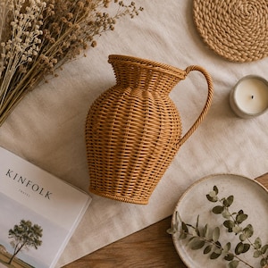May include: A light brown woven wicker pitcher with a wide, flared rim and a curved handle. The pitcher features a tight, uniform weave pattern throughout its rounded body, creating a textured surface that is both functional and decorative.